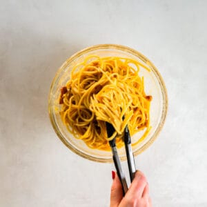 tongs tossing pasta carbonara in a glass bowl.