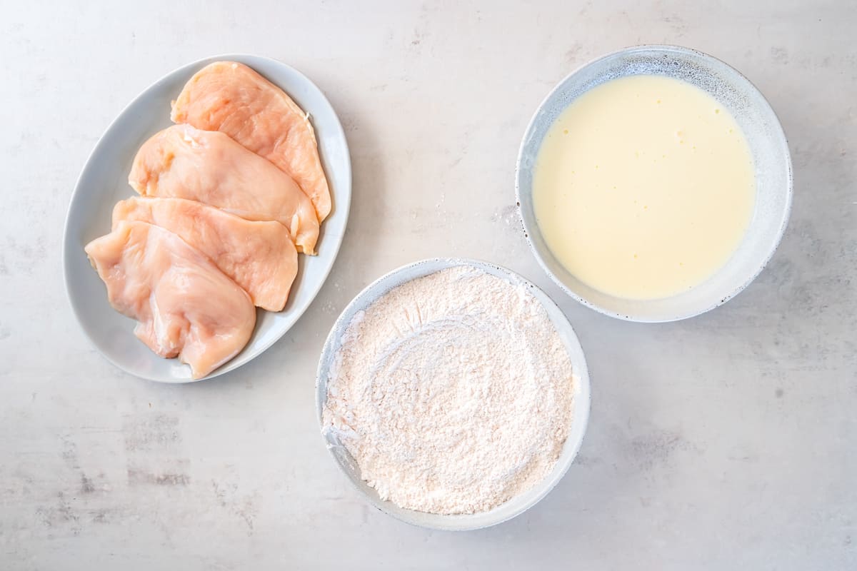 a bowl of buttermilk, a bowl of seasoned flour, and a plate of raw chicken