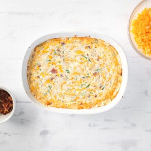 baked dip in a dish with bowls of bacon and shredded cheese next to it