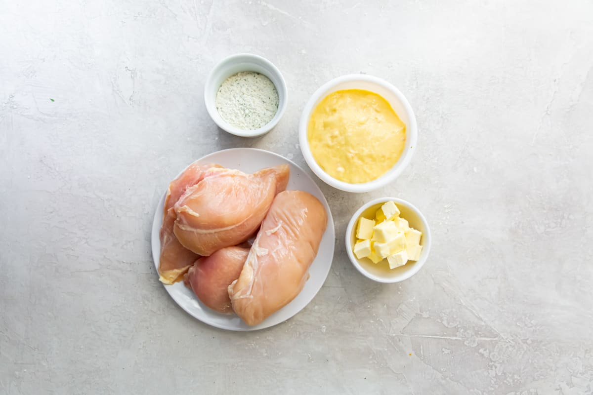 4 ingredients for Crockpot ranch chicken arranged in dishes: chicken breasts, ranch seasoning, cream of chicken soup, and cubes of butter.
