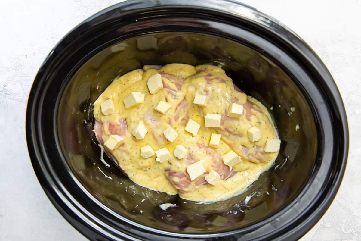 Chicken breasts, a creamy sauce, and cubes of butter in a Crockpot.