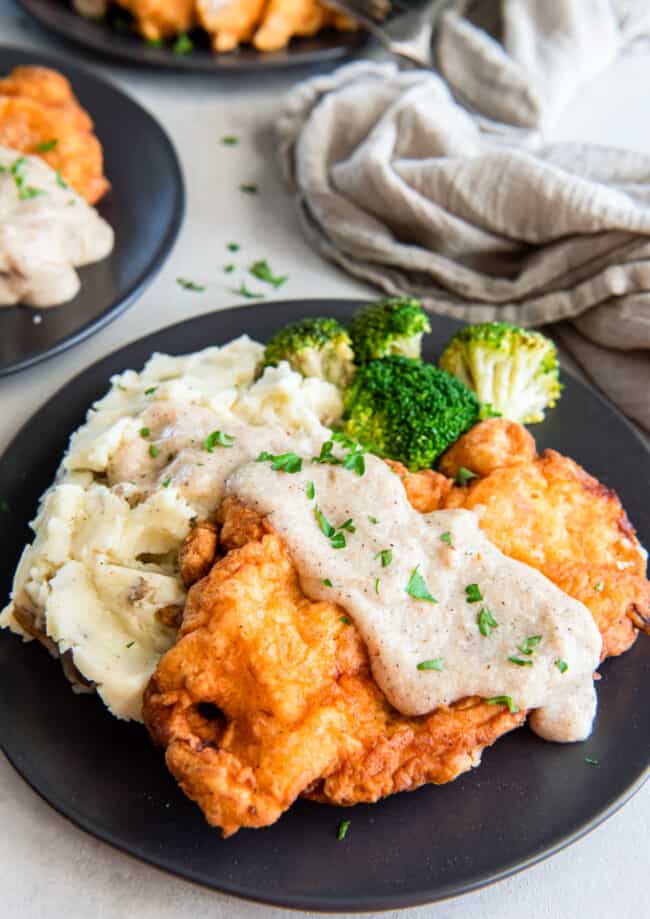 country fried chicken covered in gravy, on a plate with potatoes and broccoli