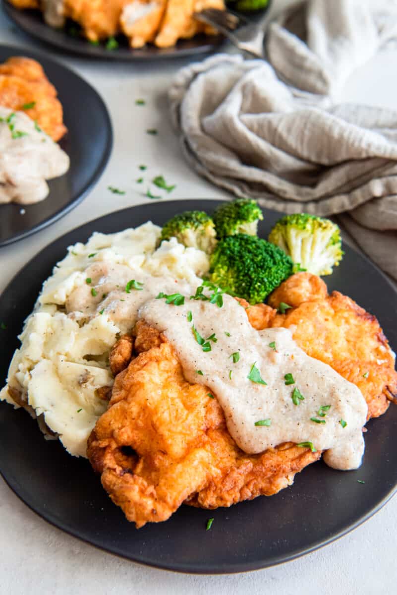 country fried chicken covered in gravy, on a plate with potatoes and broccoli