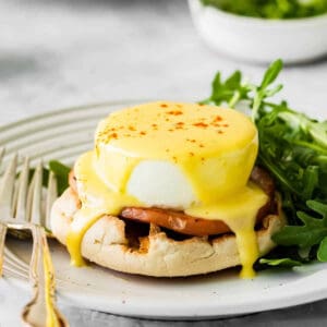 a serving of eggs benedict drenched in creamy hollandaise sauce on a white plate with a side salad and fork.