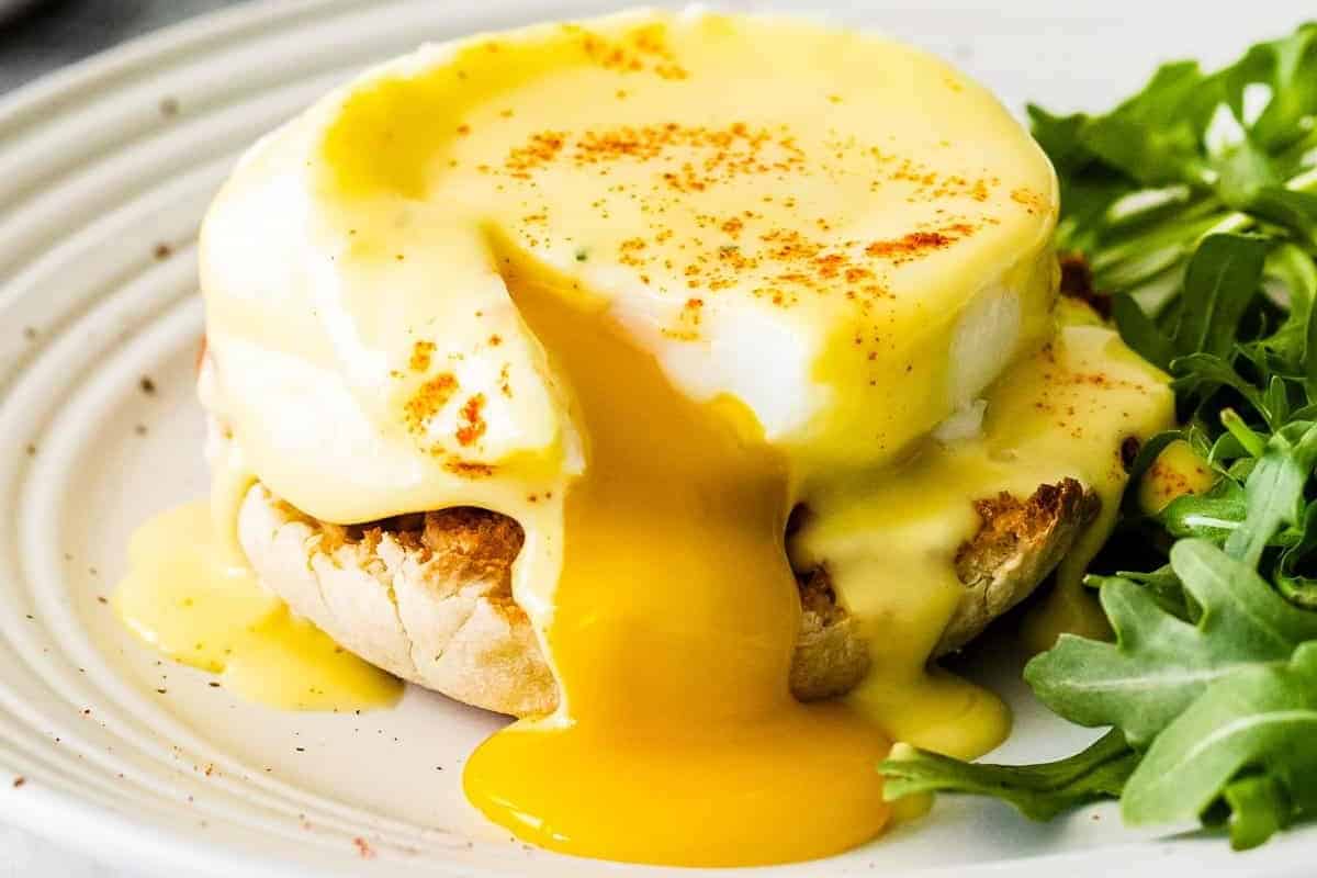 egg yolk oozing out of a cut serving of eggs benedict on a white plate with a side salad.