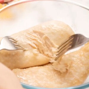 shredding cooked chicken breasts with two forks in a glass bowl.