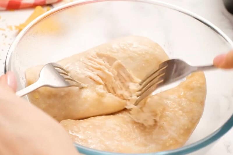 shredding cooked chicken breasts with two forks in a glass bowl.