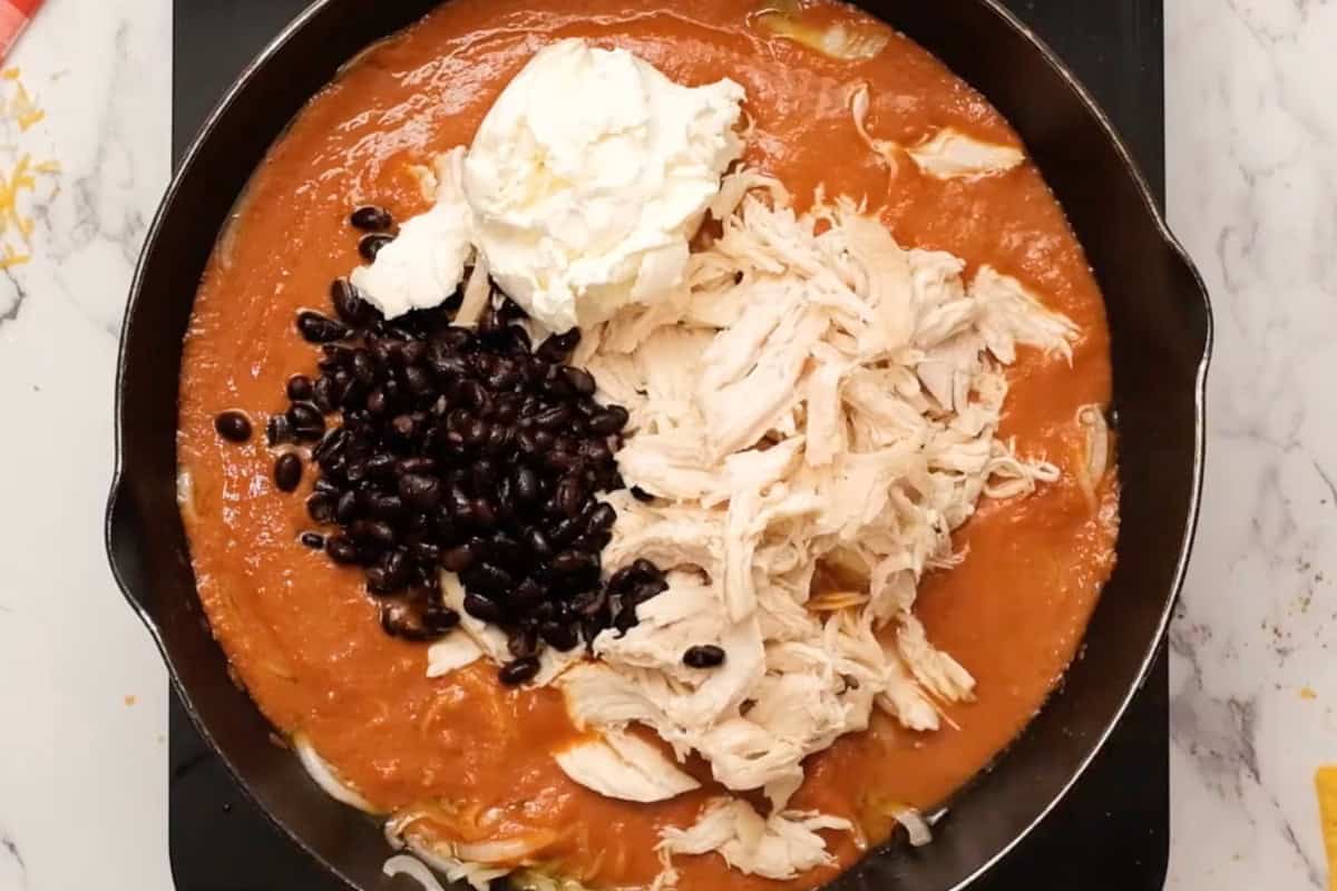adding shredded chicken, black beans, and cream cheese to enchilada sauce in a skillet.