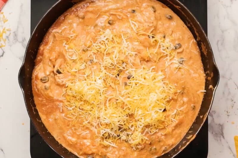adding shredded cheese to chicken enchilada dip in a skillet.
