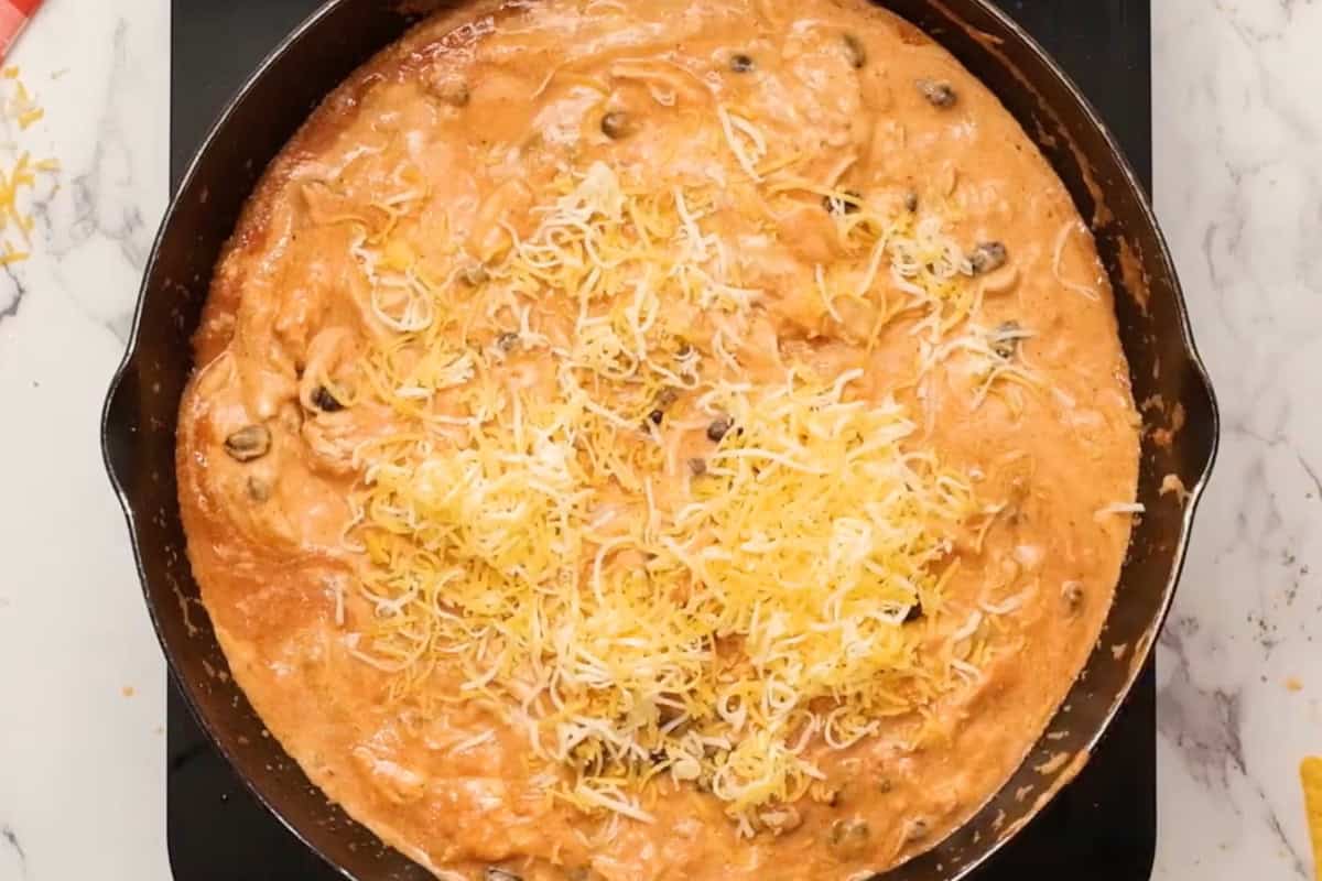 adding shredded cheese to chicken enchilada dip in a skillet.