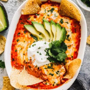 a baking dish filled with chicken enchilada dip, topped with chips, sour cream, avocado, and cilantro.