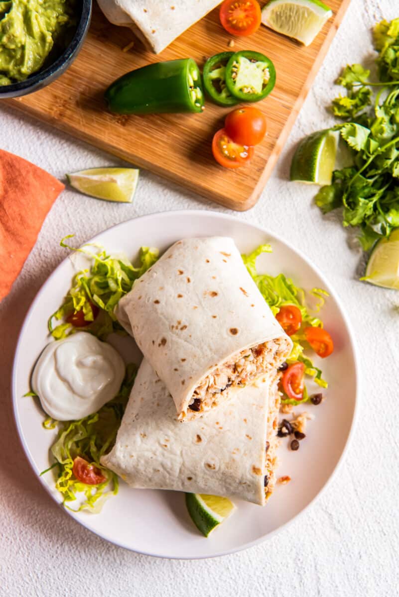 overhead view of a chicken burrito on a white plate, next to a cutting board with ingredients scattered