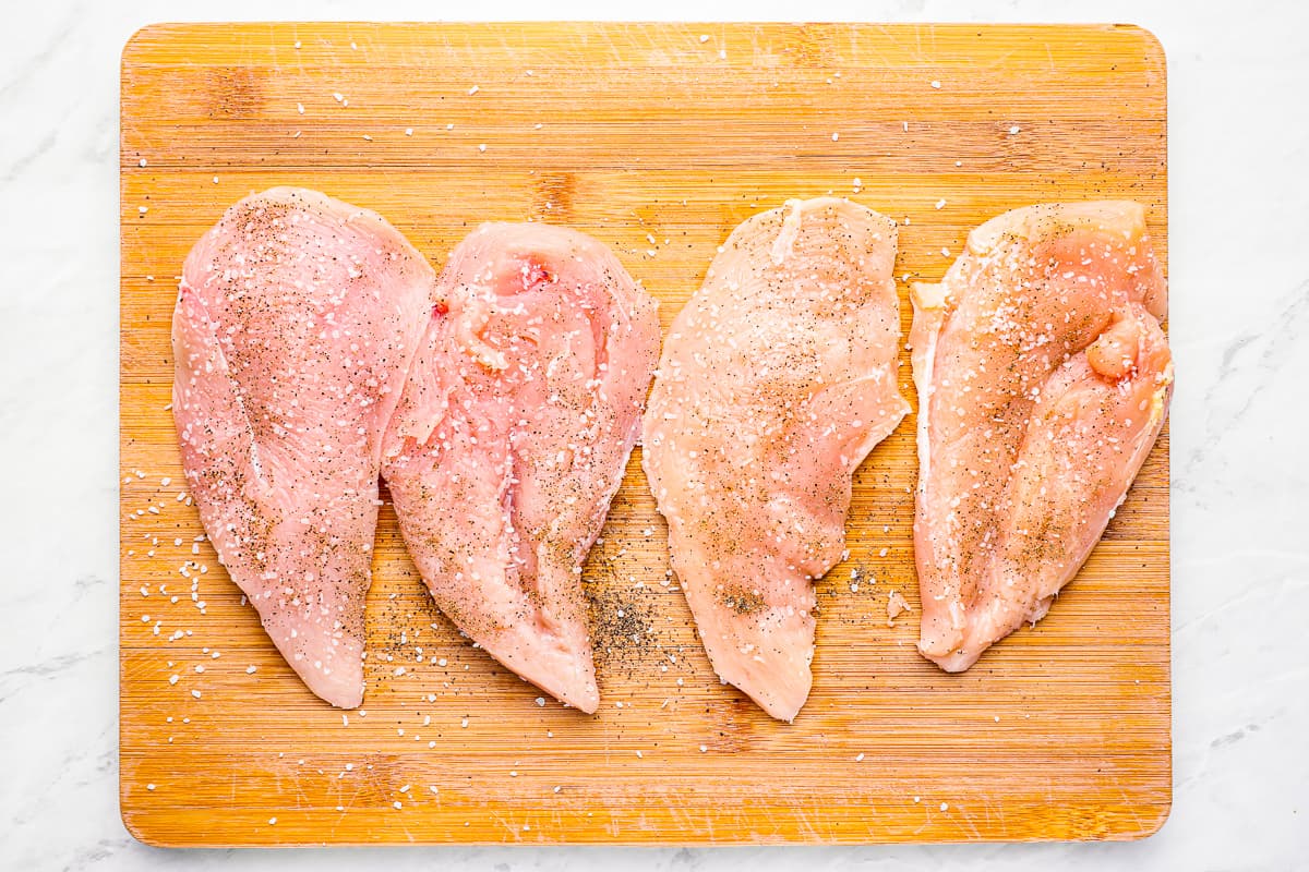 4 raw chicken breasts lined up on a cutting board, seasoned with salt and pepper.