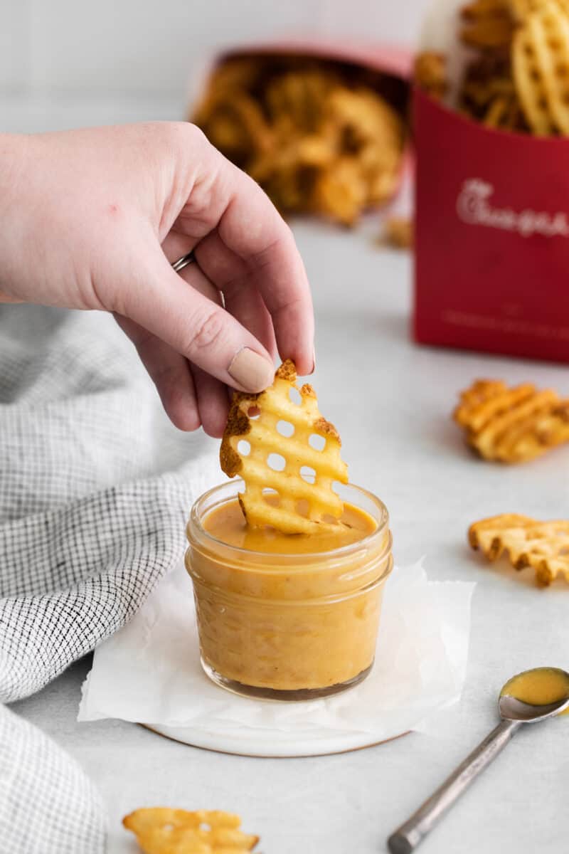 three-quarters view of a hand dipping a waffle fry into a small glass jar full of chick fil a sauce on a white napkin.