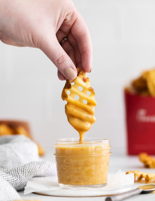 side view of a hand lifting a dipped waffle fry from a glass jar full of chick fil a sauce on a white napkin.