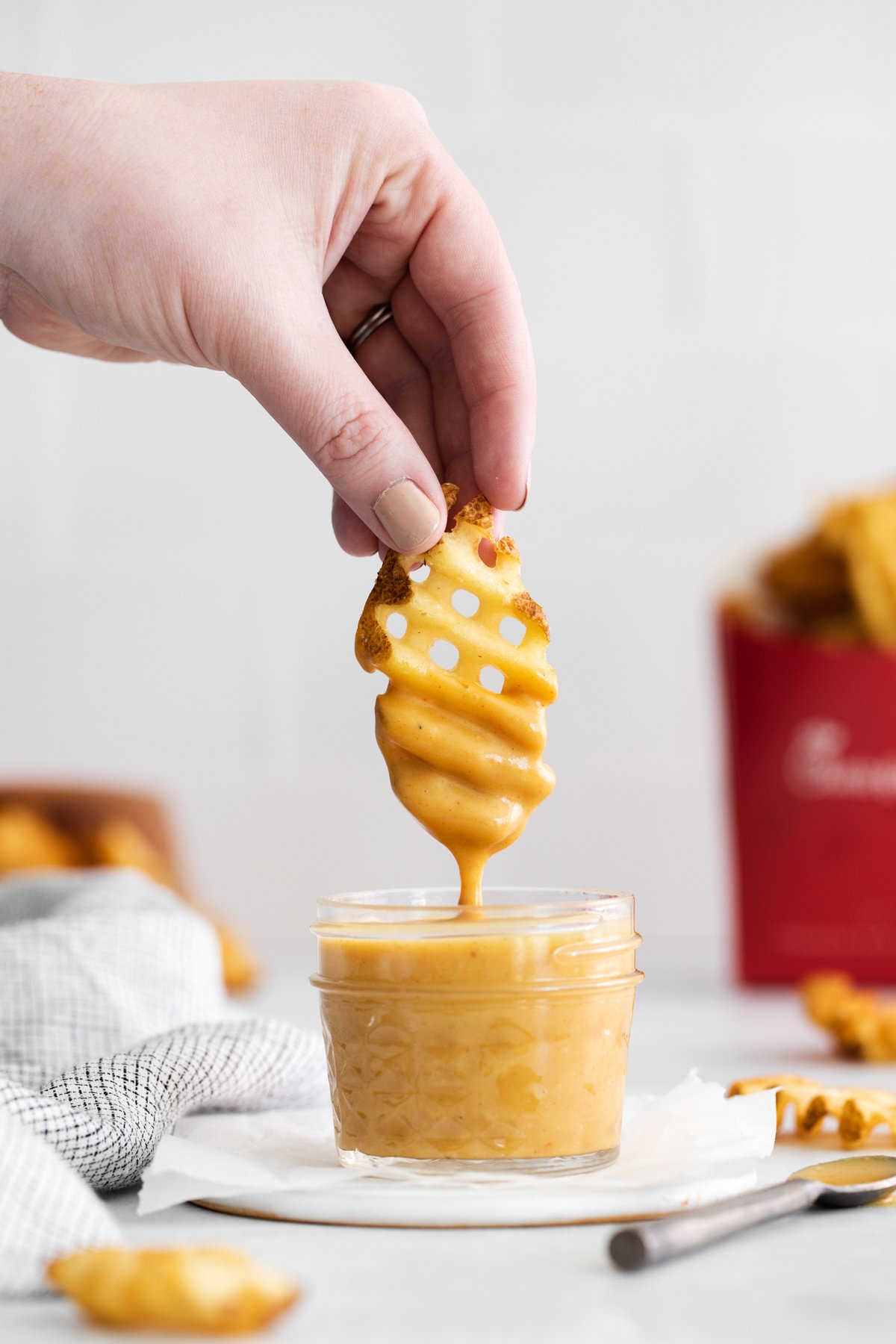 side view of a hand lifting a dipped waffle fry from a glass jar full of chick fil a sauce on a white napkin.