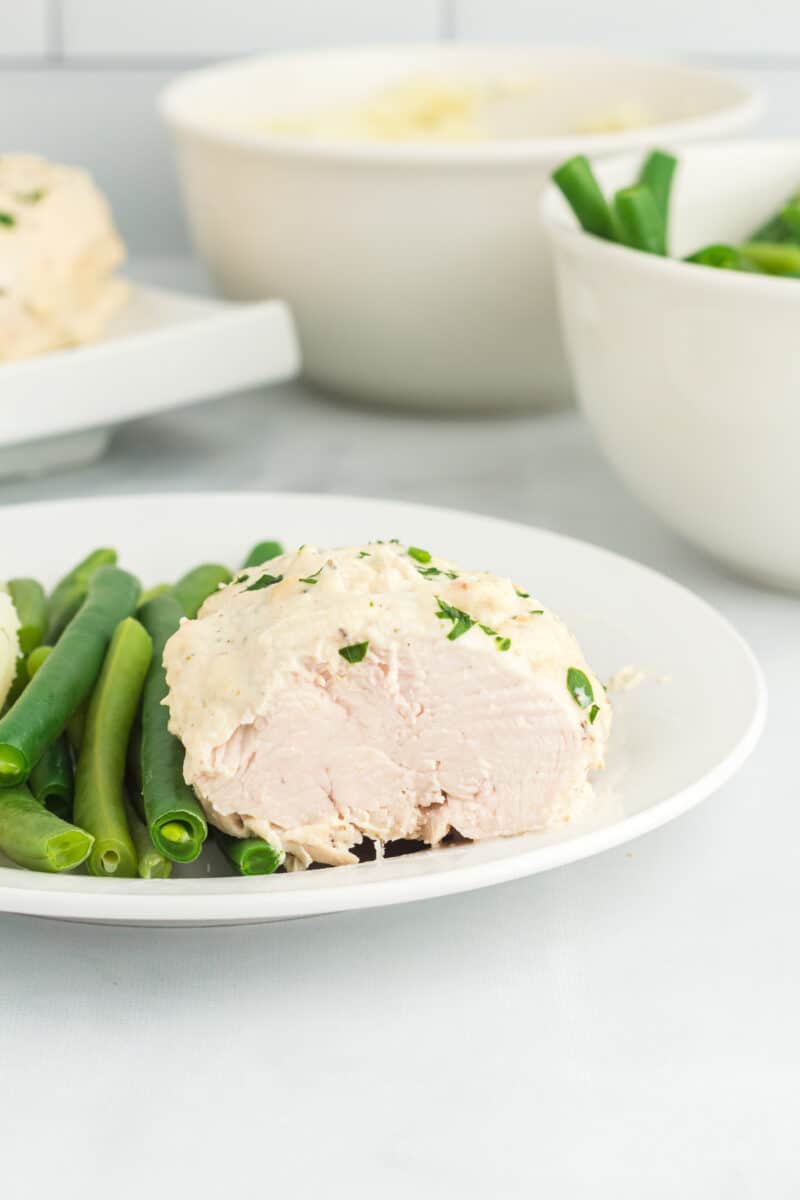 a sliced creamy herb chicken breast on a white plate with green beans.