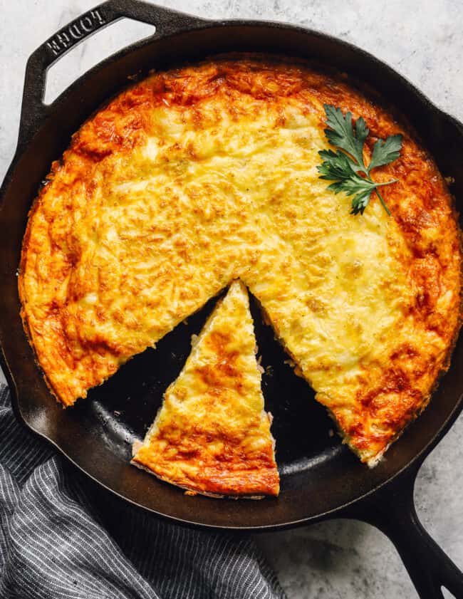 overhead view of a cut ham and cheese frittata in a cast iron pan with a small slice placed in a large gap.