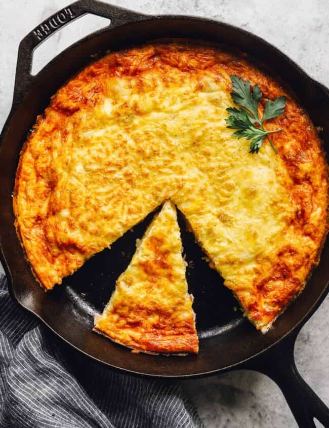 overhead view of a cut ham and cheese frittata in a cast iron pan with a small slice placed in a large gap.