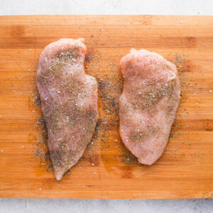 two uncooked and seasoned chicken breasts on a cutting board