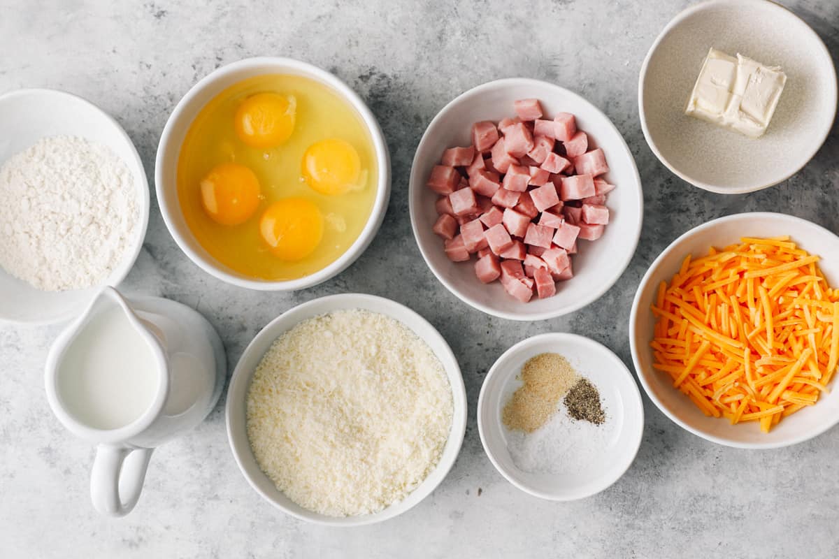Ingredients for a breakfast soufflé arranged in individual bowls: eggs, cheese, ham, butter, flour, and seasonings.