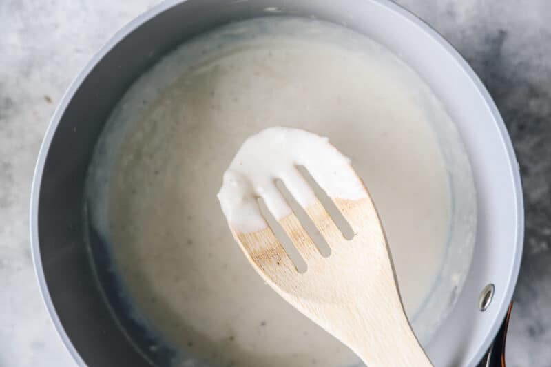 A wooden spoon is being used to stir a white sauce for a breakfast soufflé in a pan.