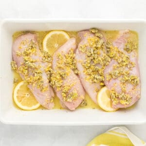4 raw chicken breasts coated in lemon butter sauce and lemon slices in a rectangular baking dish.