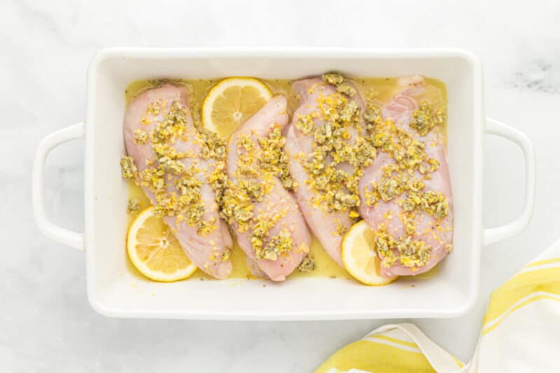 4 raw chicken breasts coated in lemon butter sauce and lemon slices in a rectangular baking dish.