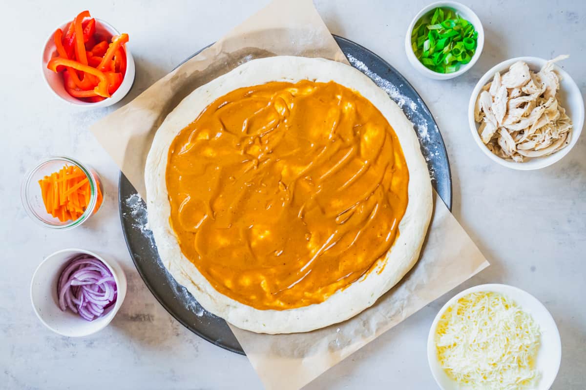 pizza dough slathered with peanut sauce, surrounded by small bowls of ingredients
