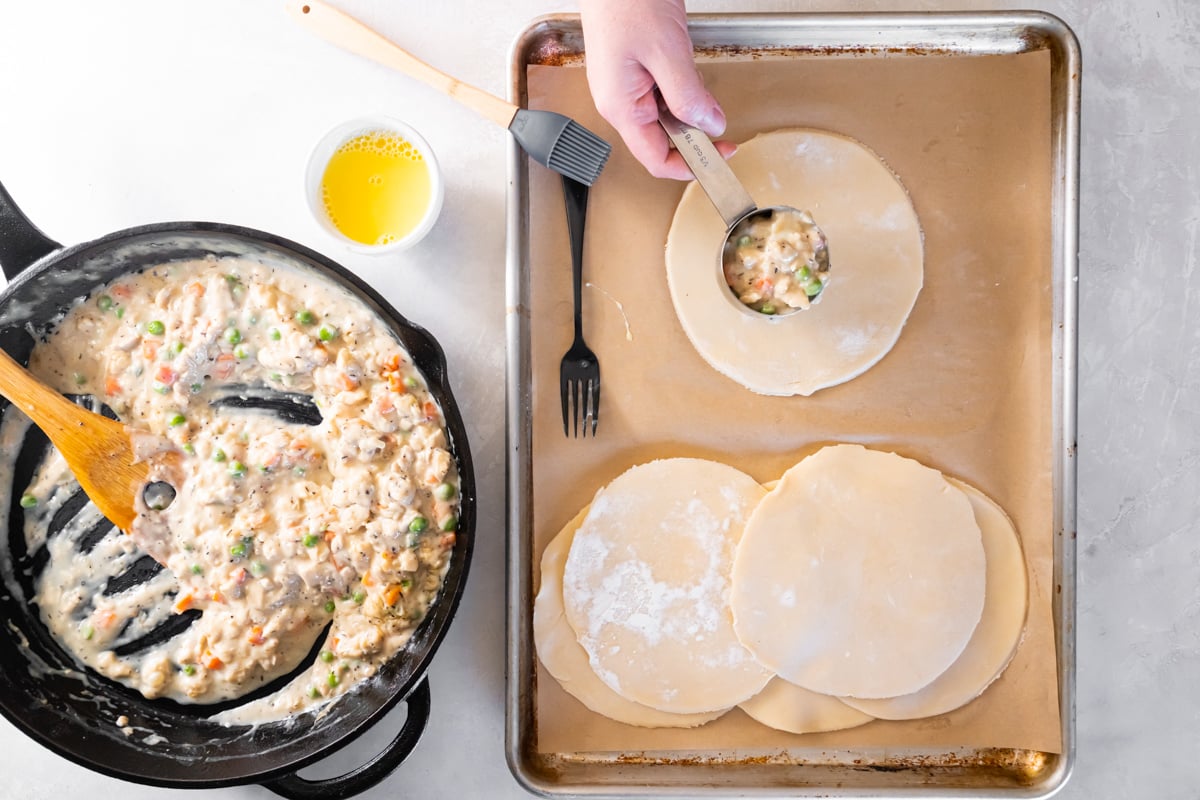 A hand using a measuring scoop to fill discs of pie dough with chicken pot pie filling.