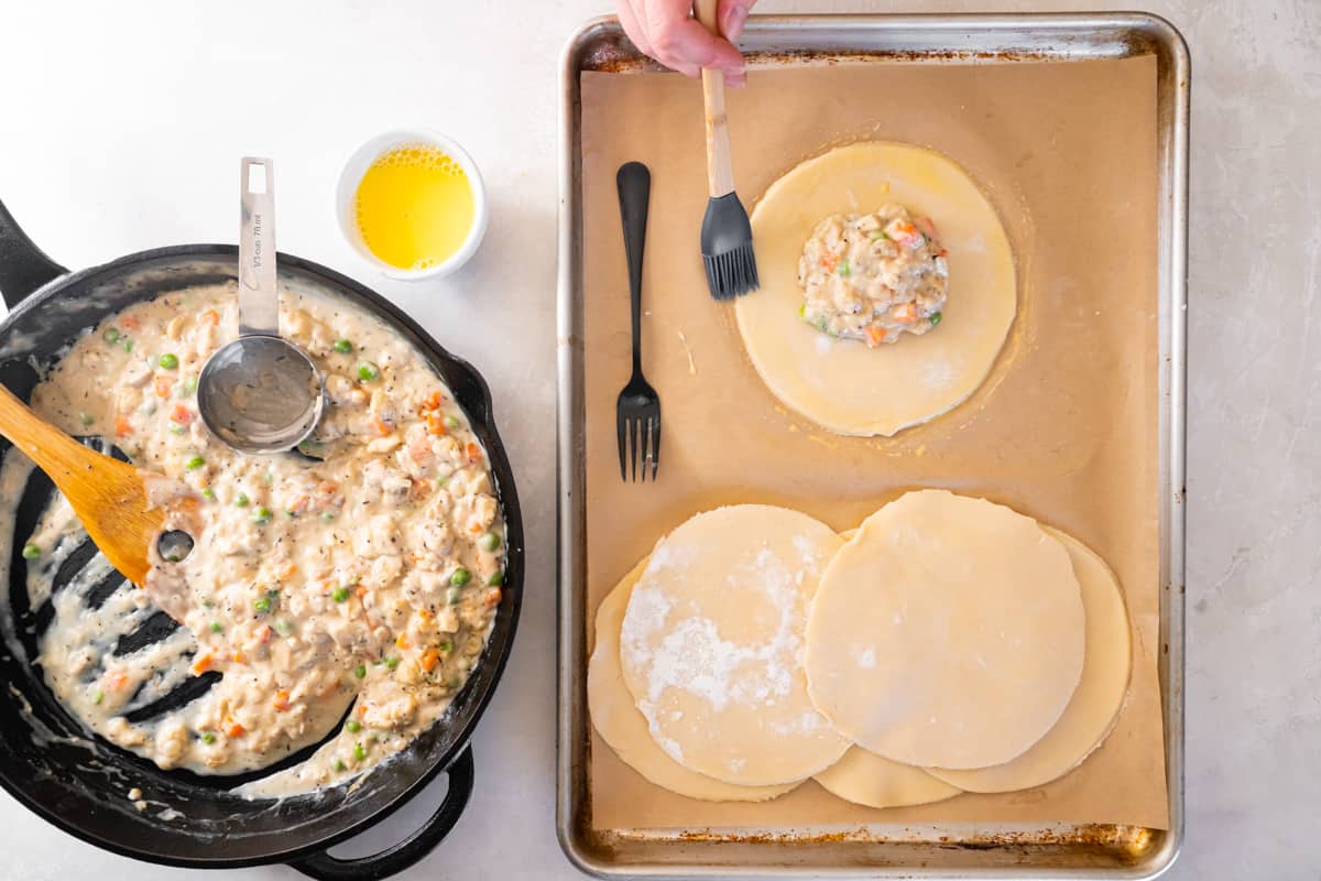 A hand using a basting brush to brush egg wash around the outside of a circle of pie dough filled with chicken filling on a baking sheet.