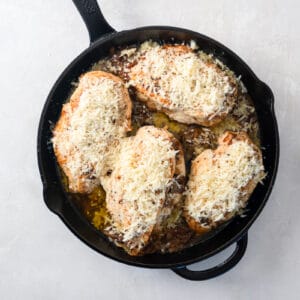 cheese sprinkled over 4 french onion stuffed chicken breasts in a cast iron skillet.