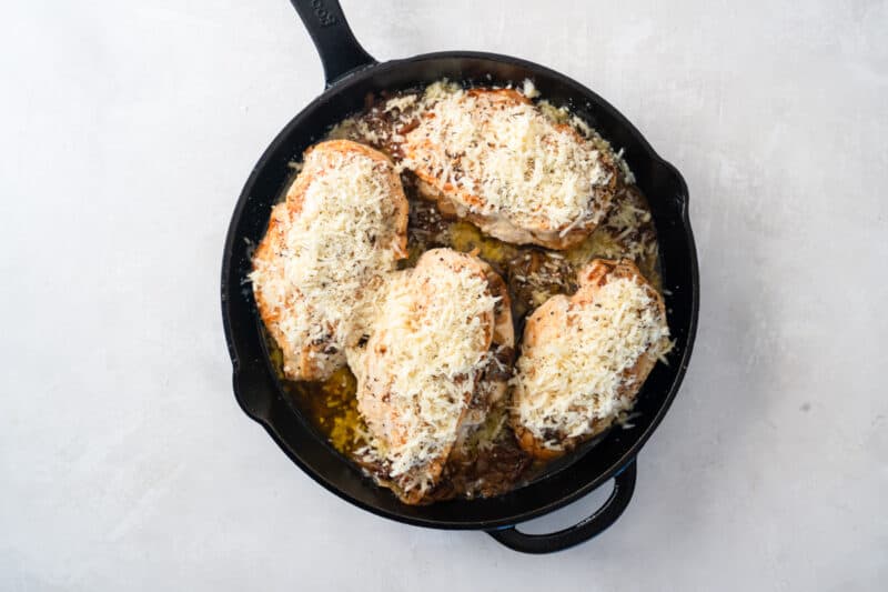 cheese sprinkled over 4 french onion stuffed chicken breasts in a cast iron skillet.