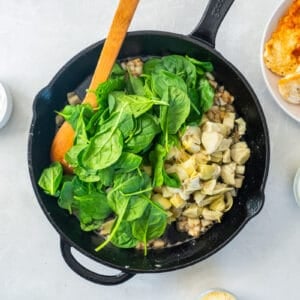 baby spinach added to spinach artichoke sauce in a cast iron skillet with a wooden spoon.