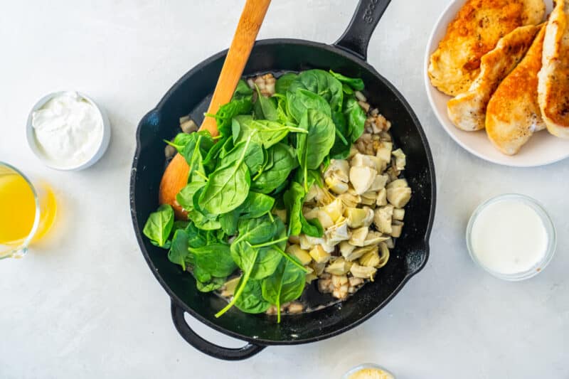 baby spinach added to spinach artichoke sauce in a cast iron skillet with a wooden spoon.