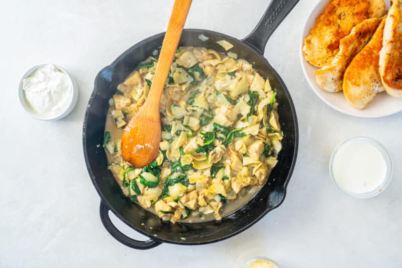A skillet full of spinach-artichoke chicken with a wooden spoon.
