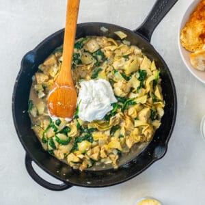 sour cream added to spinach artichoke sauce in a cast iron skillet with a wooden spoon.