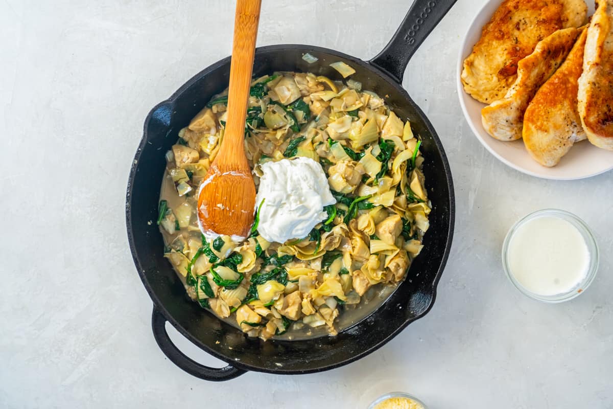 sour cream added to spinach artichoke sauce in a cast iron skillet with a wooden spoon.