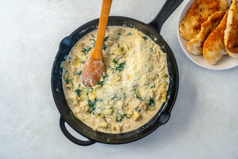 parmesan cheese added to spinach artichoke sauce in a cast iron skillet with a wooden spoon.