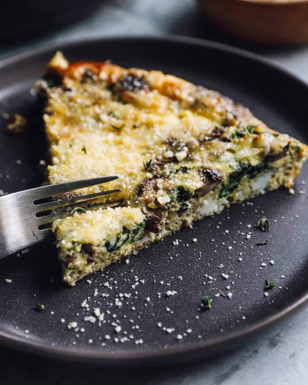 using a fork to cut a slice of mushroom frittata on a black plate.