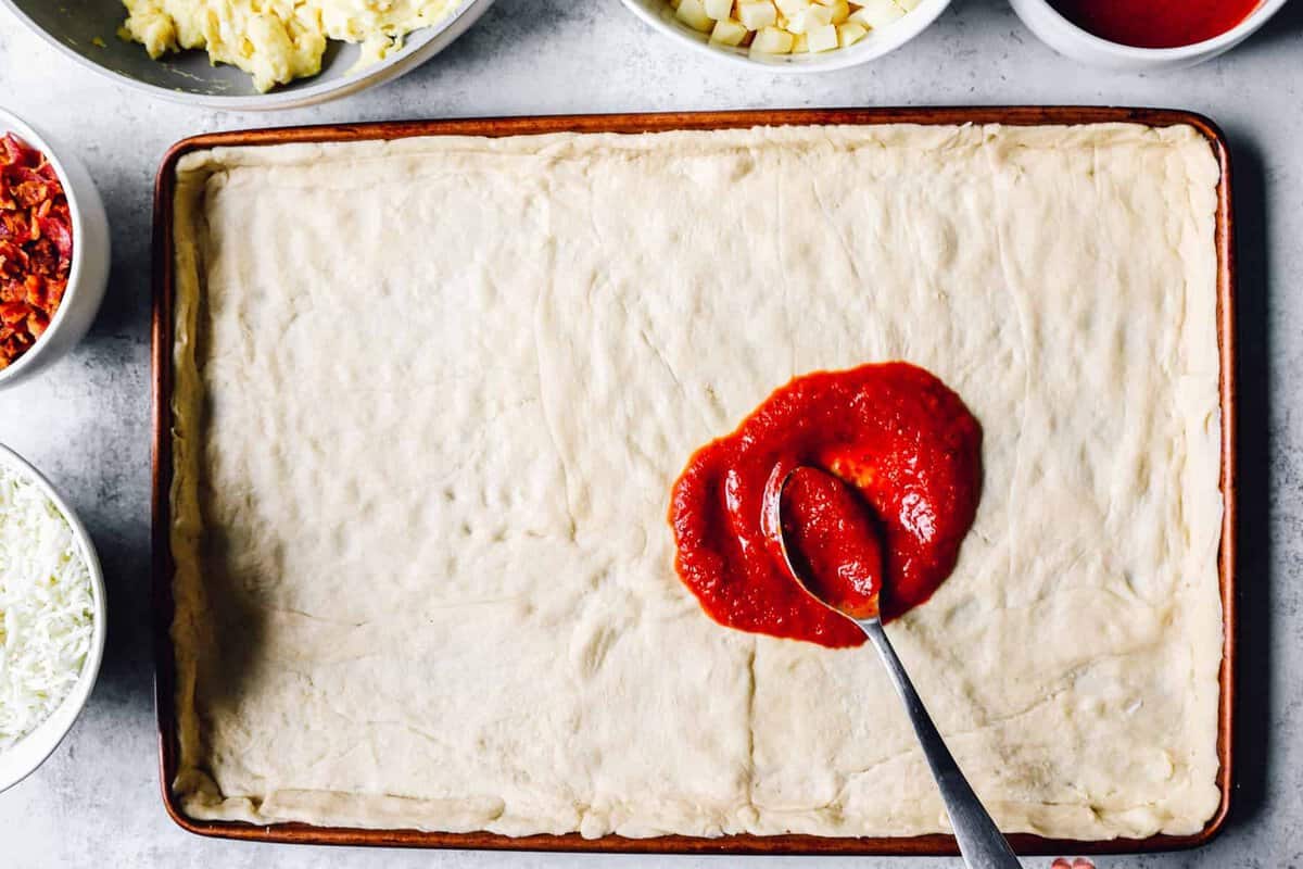 pizza sauce spread on crescent roll crust in a sheet pan.