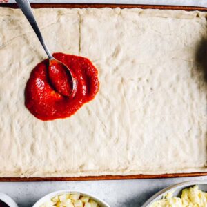 pizza sauce spread on crescent roll crust in a sheet pan.