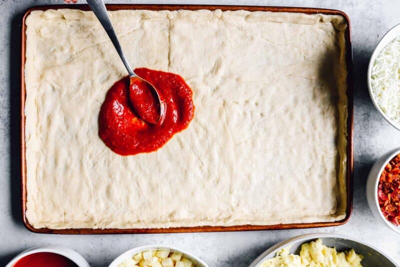 pizza sauce spread on crescent roll crust in a sheet pan.
