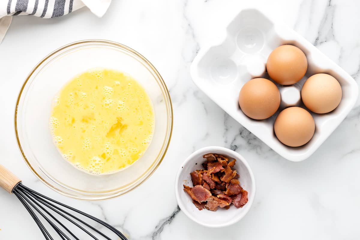 Beaten eggs in a glass bowl, next to a bowl of bacon crumbles and a dish with whole eggs.