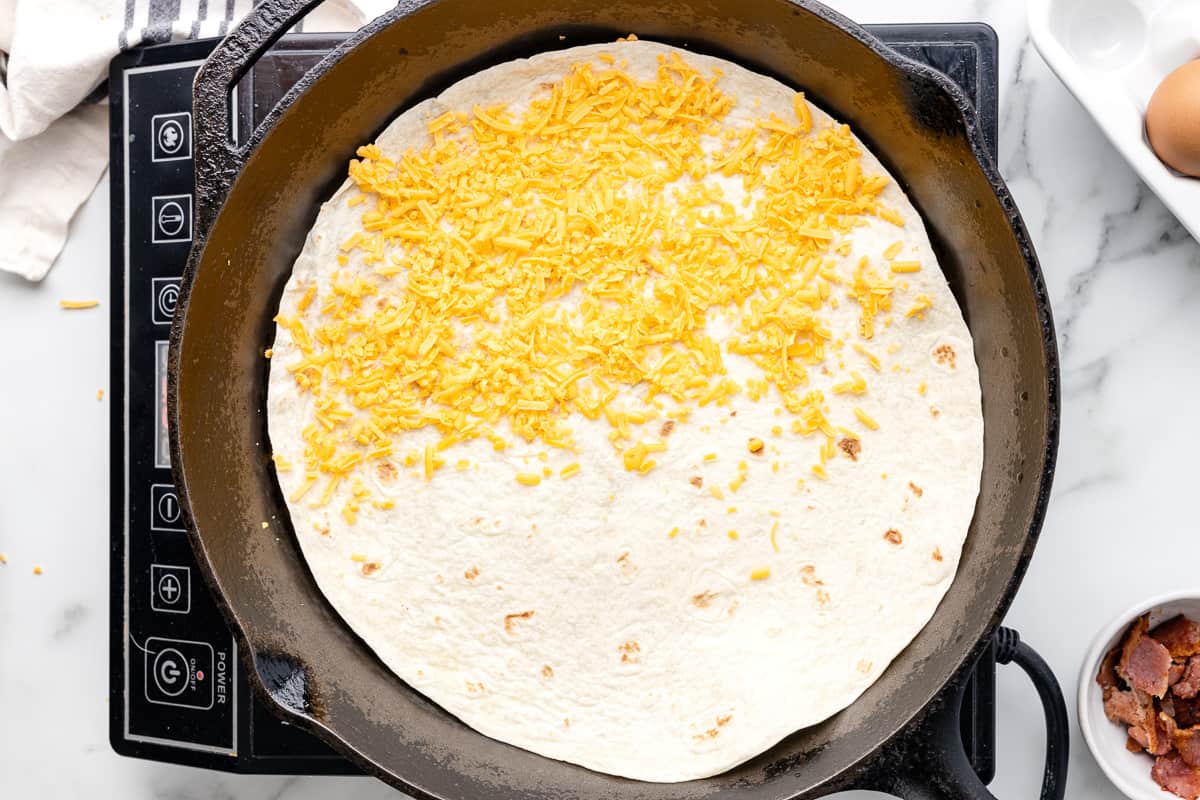 Shredded cheese sprinkled over half of a tortilla in a pan.