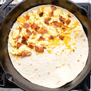 egg, bacon, and cheese mixture spread over half of a tortilla in a pan.