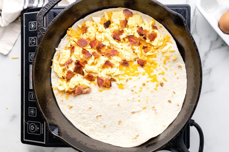 egg, bacon, and cheese mixture spread over half of a tortilla in a pan.