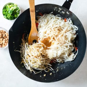 rice noodles added to chicken pad thai in a wok with a wooden spoon.