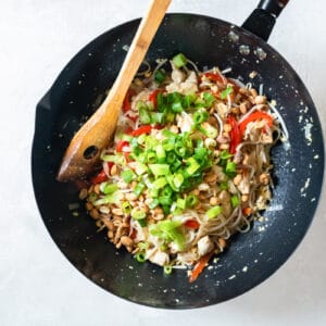 green onion added to chicken pad thai in a wok with a wooden spoon.