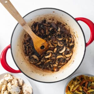 cooked mushrooms and veggies in a red dutch oven with a wooden spoon.